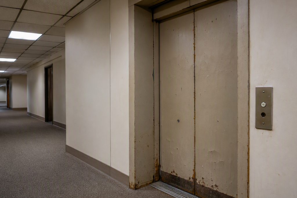 Worn, aging commercial elevator with dented steel doors, corroded threshold, and outdated call button panel — common end-of-life warning signs in a Phoenix AZ office building.